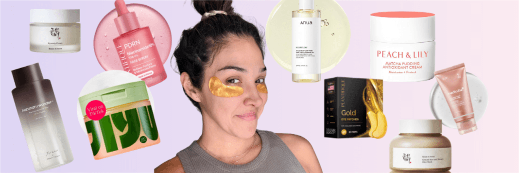 A woman with dark hair tied in a bun wears a gray sleeveless top and gold under-eye patches. Various skincare products, including jars, serums, creams, and face masks, surround her against a light purple and pink background.
