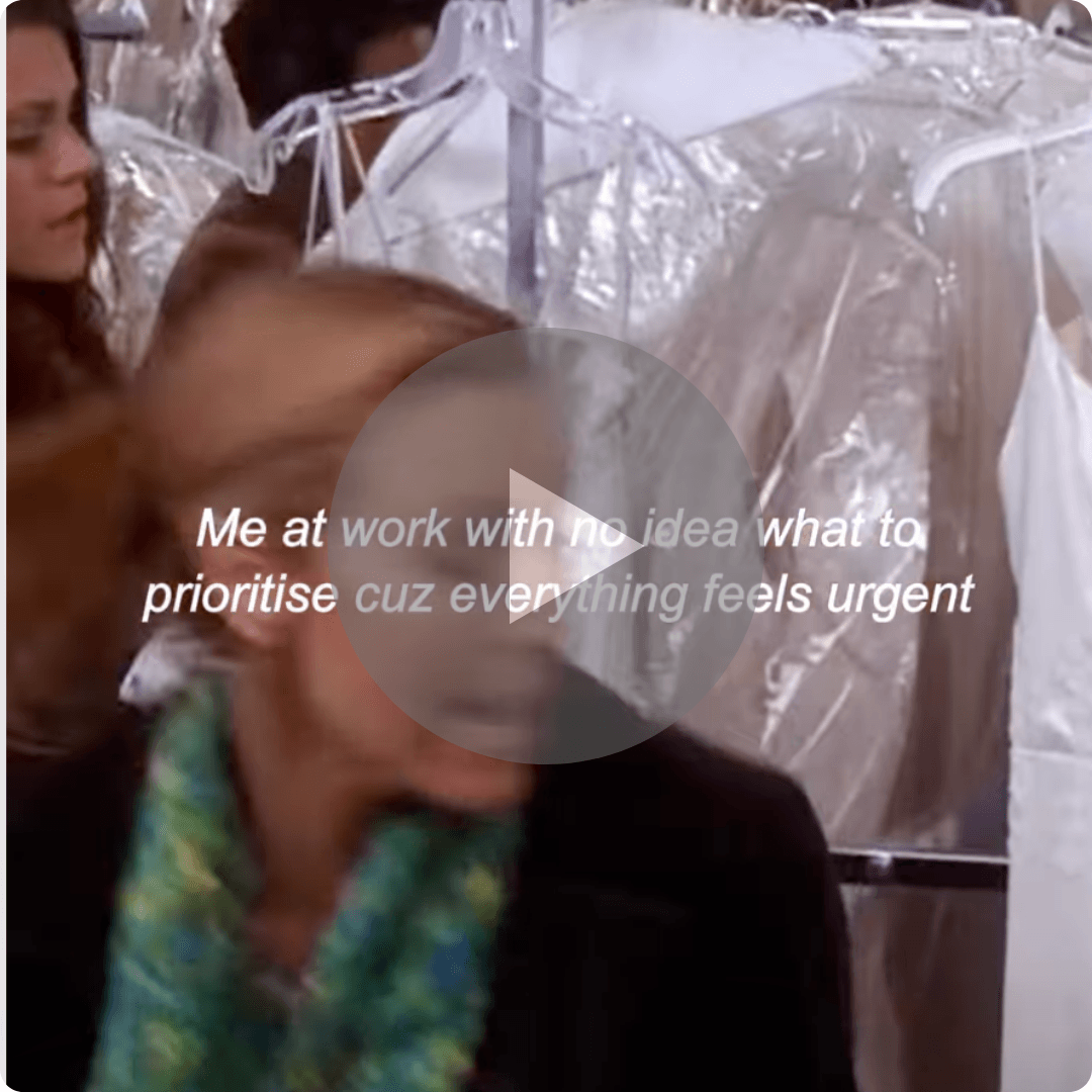 A video thumbnail features a woman in a green patterned top standing in front of clothing racks covered in clear plastic. White text across the center reads, “Me at work with no idea what to prioritise cuz everything feels urgent,” with a large play button overlay.