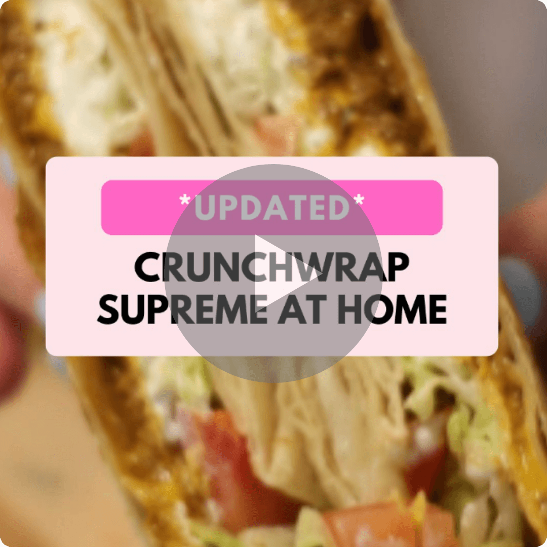 A close-up of a folded, golden-brown Crunchwrap filled with meat, lettuce, and other ingredients fills the background. A light pink text box in the center reads “UPDATED CRUNCHWRAP SUPREME AT HOME,” with a large translucent play button over it.